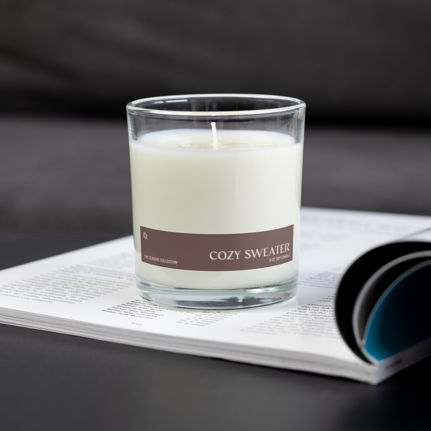 Candle labeled 'Cozy Sweater' on an open magazine with a dark background