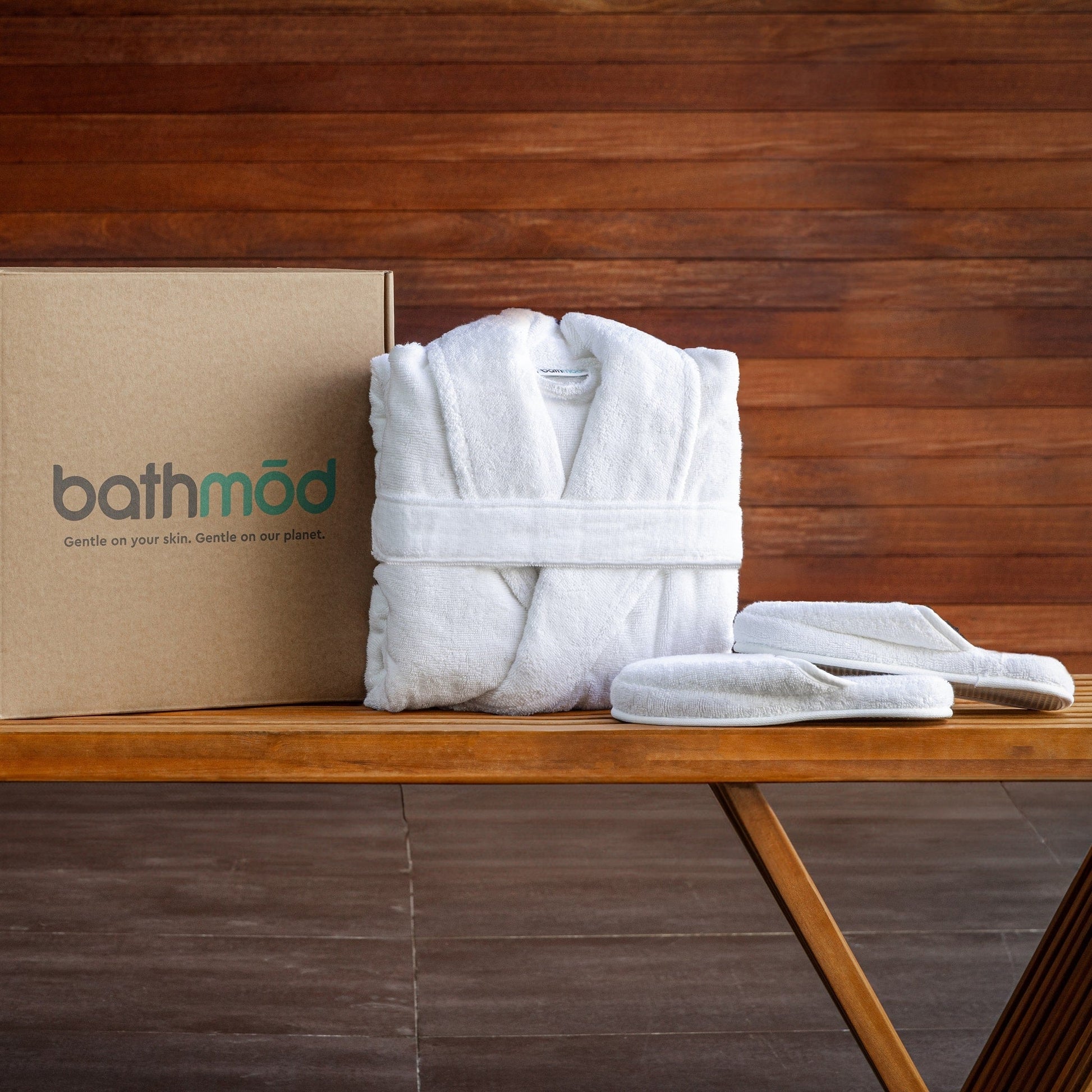 White bathrobe and slippers on a wooden table with a 'bathmōd' box in the background.